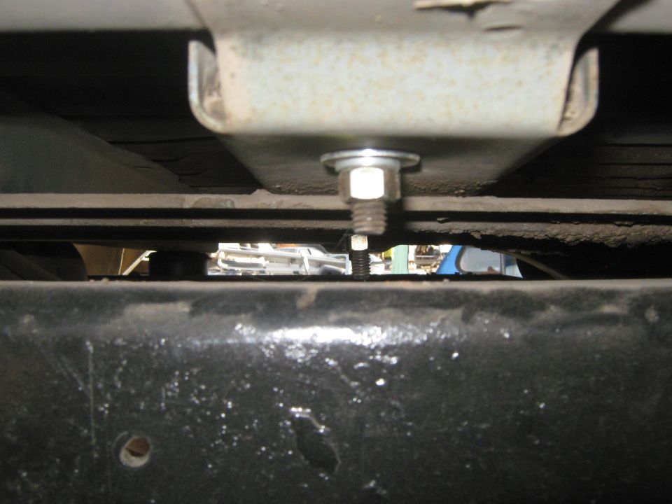 Underside view of a vehicle frame showing a bolt and nut assembly. Other vehicles are visible in the background
