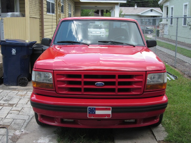 Red 1992 Ford F-150 truck front view, parked in driveway. Another white truck is visible in the background