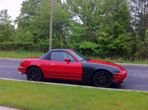 1990 Mazda Miata - A red convertible with a black hood sits parked on the grass next to a road.
