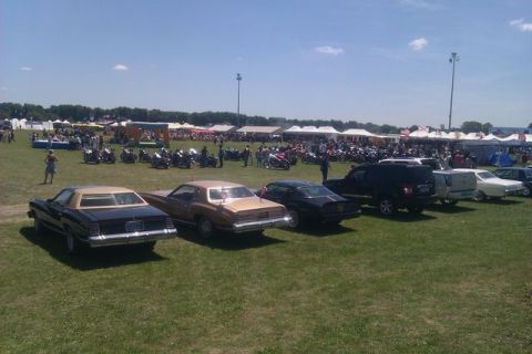 1991 Ford LTD Crown Victoria - An outdoor event features a variety of cars and motorcycles parked on a grassy field under a clear sky.