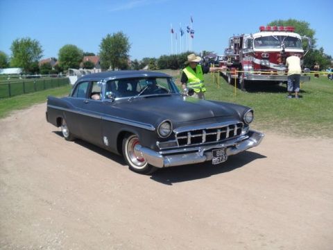 1991 Ford LTD Crown Victoria - A dark gray 1955 Chrysler New Yorker sedan is parked on a dirt road with a fire truck in the background.