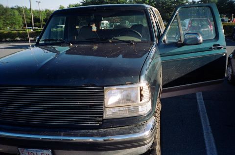 1994 Ford Bronco - Dark green SUV parked in a lot with the driver's door ajar, showing the front grille and headlight.