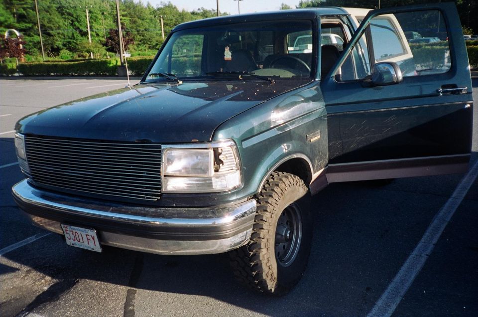 Dark green Ford Bronco with tan top, open driver's door, billet grille and mud tires in a lot