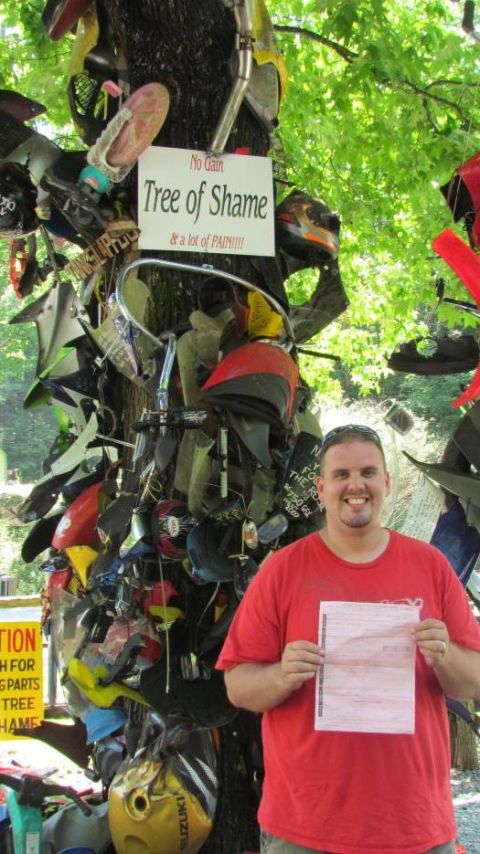 2007 Ford Mustang - A man smiles while holding a document in front of a tree covered in motorcycle parts, a testament to past crashes.