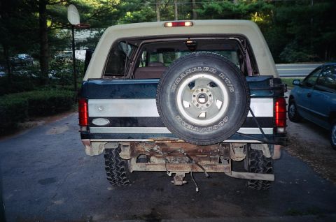 1994 Ford Bronco - Rear view of a Ford Bronco with a spare tire mounted, showing its two-tone paint and a rusty bumper.