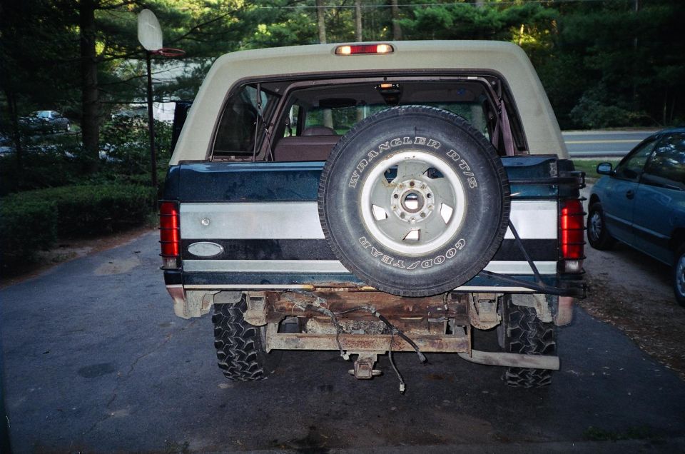 Ford Bronco rear with spare tire, dark green and tan body, rusty bumper, and a blue car parked nearby