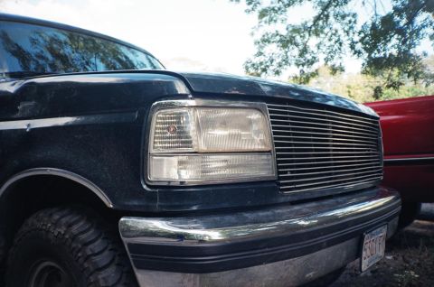 1994 Ford Bronco - Close-up of a Ford Bronco's front end, showing the grille and headlight assembly. Another vehicle is partially visible.