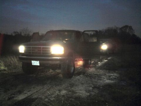 1994 Ford Bronco - A dark 1992 Ford F-Series truck sits on a muddy surface at night with its headlights illuminated and the driver's door ajar.