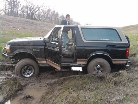 1994 Ford Bronco - A black Ford Bronco with a tan top is stuck in thick mud, with its driver's door open and a person visible inside.
