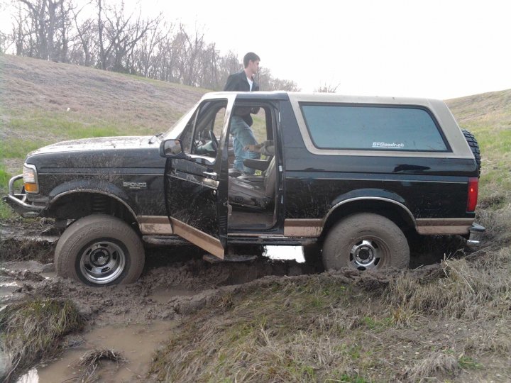 Black Ford Bronco with tan top stuck in mud, driver's door open, person in driver's seat