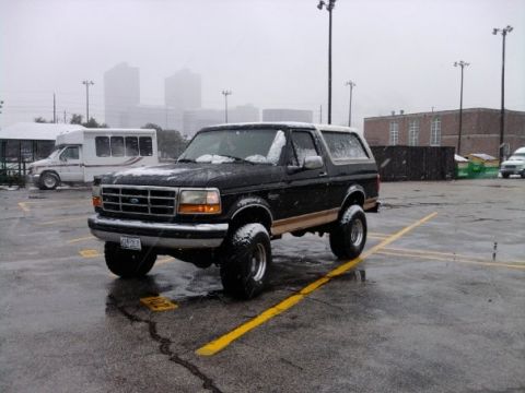 1994 Ford Bronco - A black 1994 Ford Bronco with tan trim is parked in a snowy lot, with a bus visible in the background.
