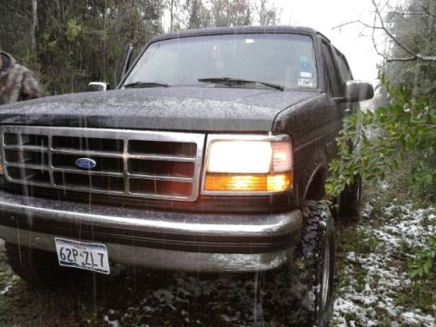 1994 Ford Bronco - A 1994 Ford Bronco sits on a muddy trail during a light snowfall, its headlights illuminated.