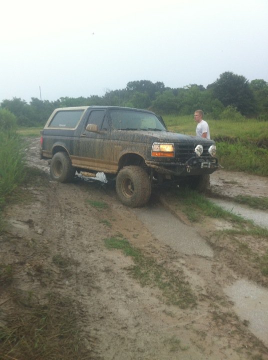 Muddy 1994 Ford Bronco driving on a dirt track with a person standing nearby