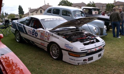 1990 Ford Probe - A white race car with numerous sponsor decals and the number 86 is displayed with its hood open, alongside other vehicles.