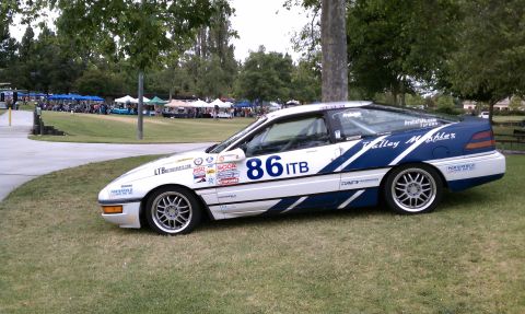 1990 Ford Probe - A white and blue 1990 Ford Probe race car with various decals is parked on a grassy area with tents and trees in the background.