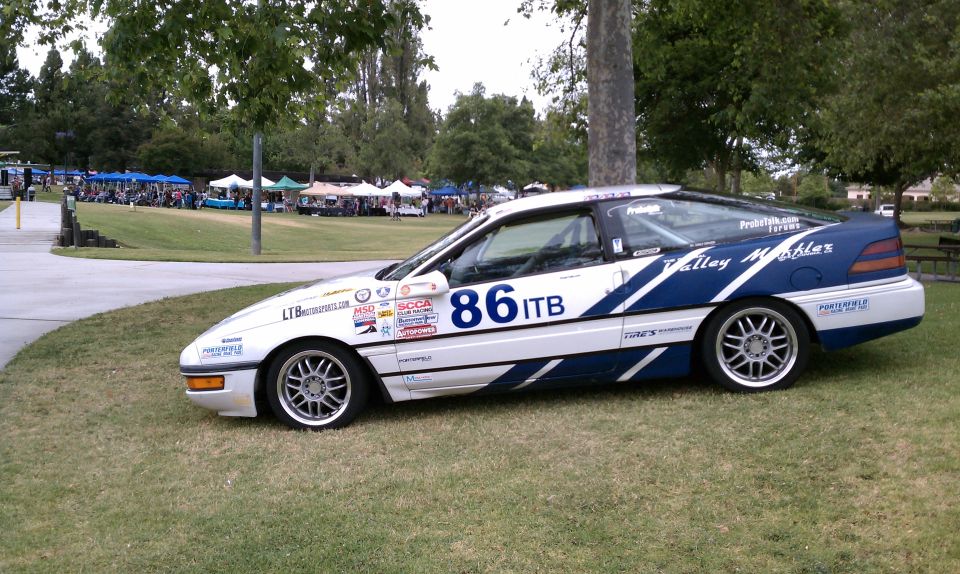 White and blue 1990 Ford Probe race car with number 86 ITB, parked on grass