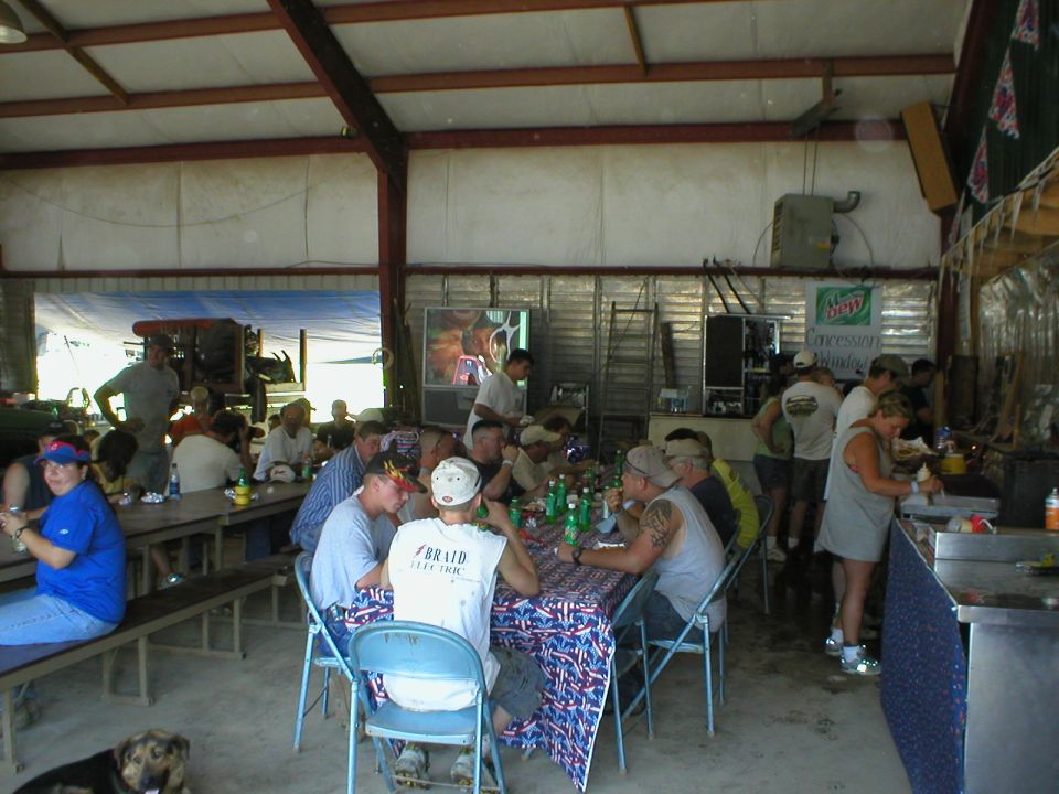 People seated at long folding tables eating under an open metal pavilion, dog lying on floor