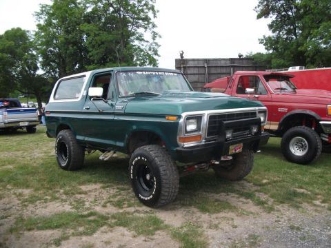 1979 Ford Bronco - A green Ford Bronco is parked on grass next to a red pickup truck, with trees and a wooden fence in the background.
