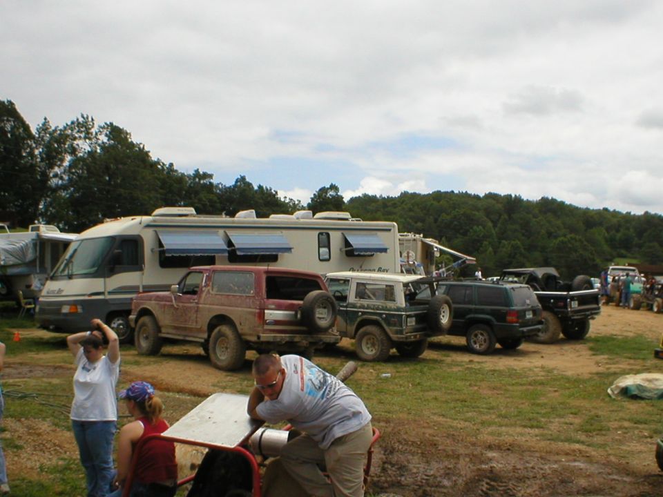 Muddy SUVs and trucks parked beside an older motorhome at an off-road event, people in foreground