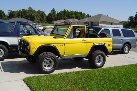 1974 Ford Bronco - A bright yellow 1974 Ford Bronco is parked on a driveway, with other vehicles visible in the background.
