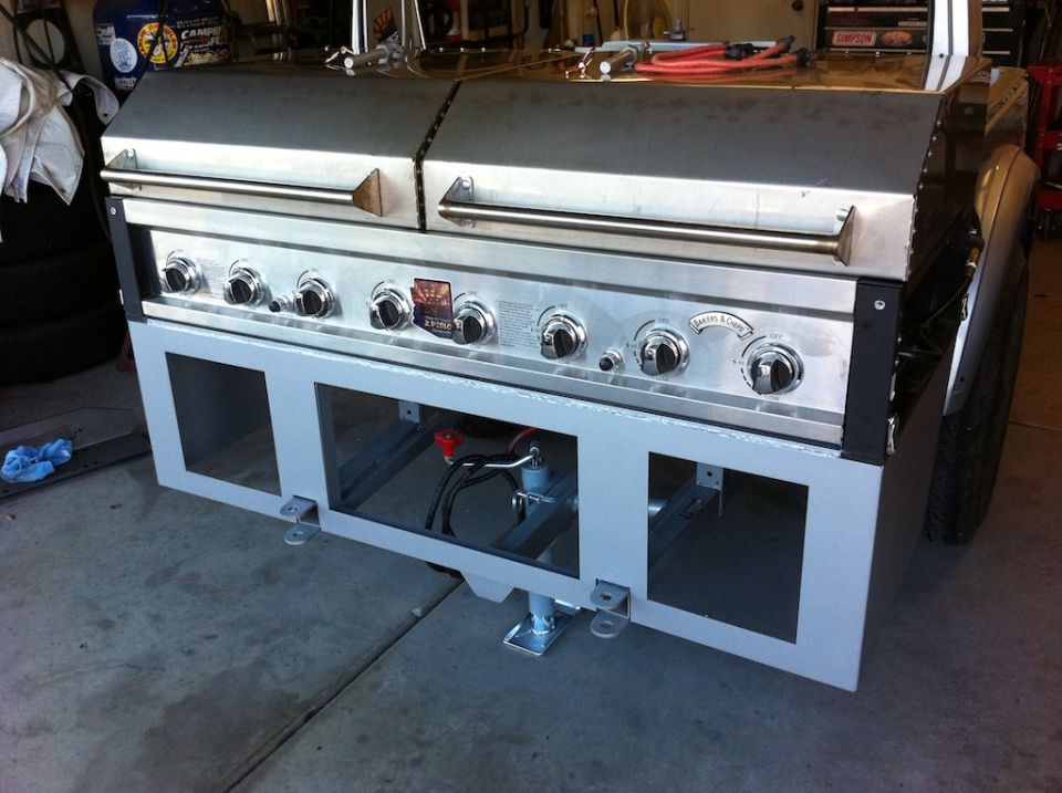 Custom grill with six burners and stainless steel lid, mounted on a vehicle frame with a visible tire and fender