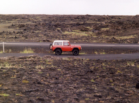 1974 Ford Bronco - A red and white 1974 Ford Bronco travels down a paved road through a barren, rocky terrain.