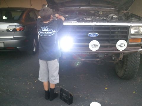1982 Ford Bronco - Boy works on the engine of a 1980 Ford Bronco, with another car nearby. The Bronco has Hella lights on its bumper.