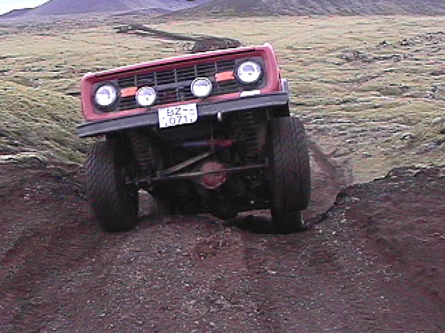 Red early Ford Bronco climbing dirt berm off-road, front axle and driving lights visible