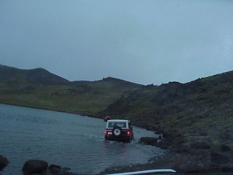 1974 Ford Bronco - Multiple off-road vehicles navigate a watery path beside a lake, with a mountainous landscape under an overcast sky.