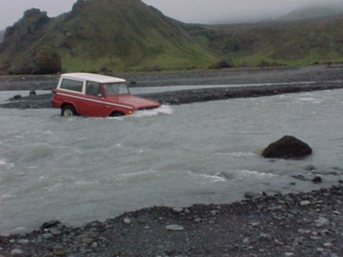 1974 Ford Bronco - A red and white SUV navigates a rocky riverbed under a cloudy sky.