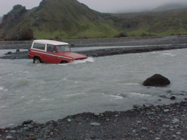 Red early Ford Bronco with white hardtop fording a wide silty river below volcanic hills