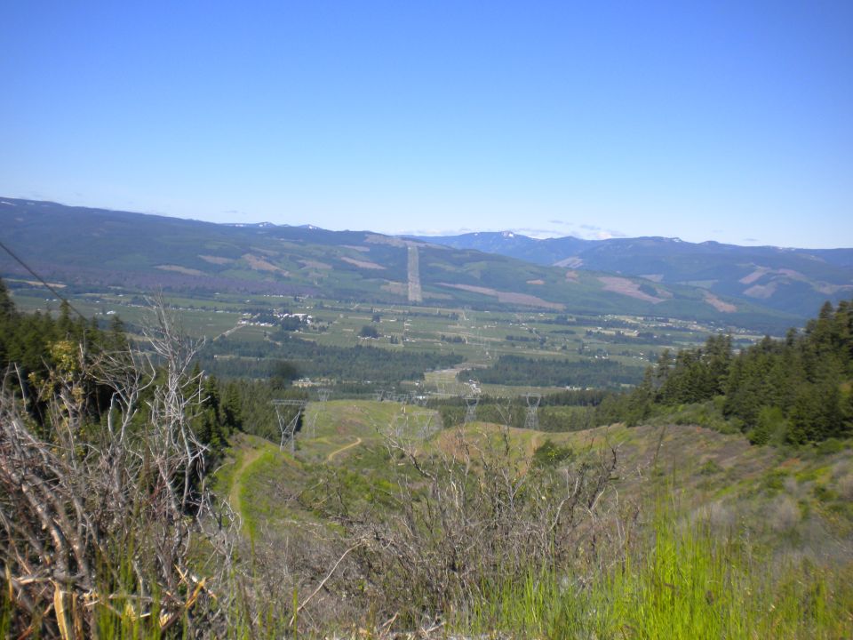 View from a hillside overlooking a wide valley with power lines and distant mountains under a clear blue sky