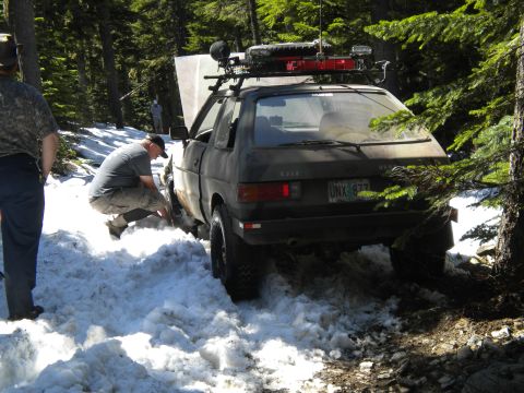 2005 Jeep Wrangler - A man works on a Subaru hatchback stuck in snow on a wooded trail, its hood open and roof rack laden with equipment.