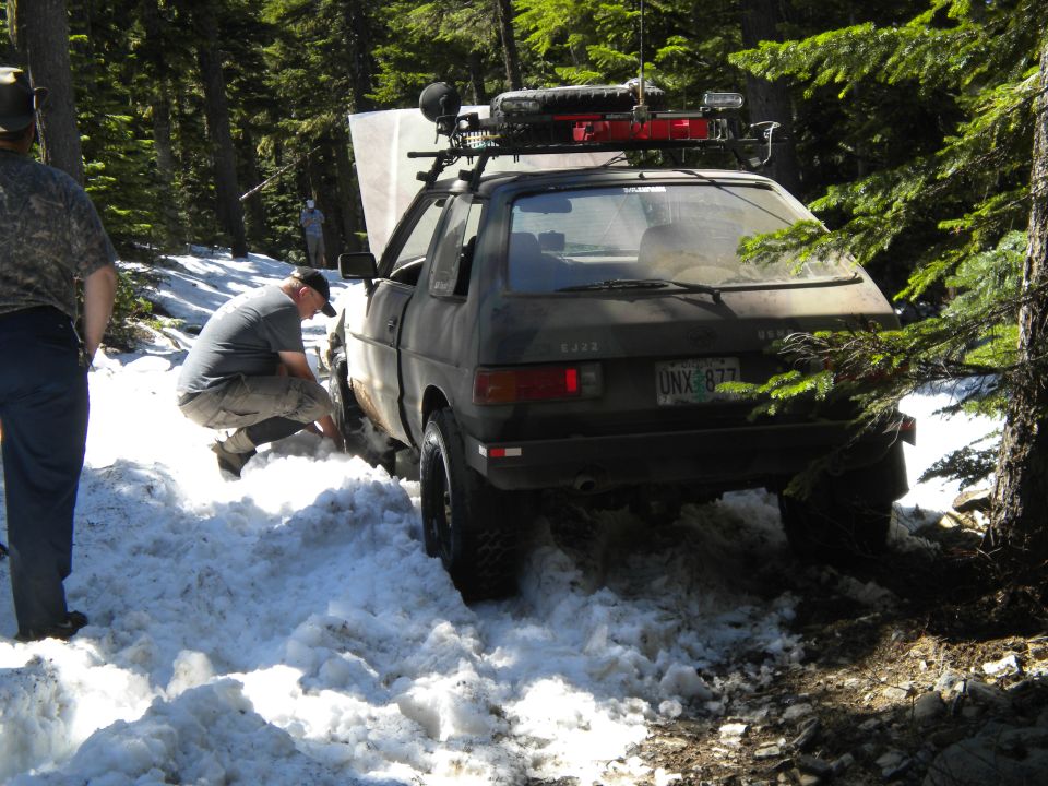 Dark green Subaru hatchback stuck in snow, hood open, with roof rack loaded with gear