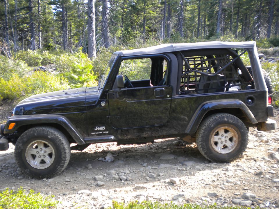 Black 2005 Jeep Wrangler Unlimited with a soft top and roll cage, parked on a rocky dirt road in a forest