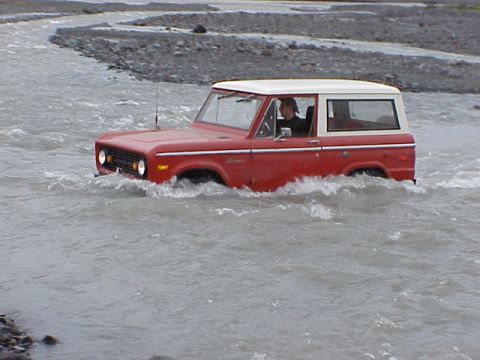 1974 Ford Bronco - A red 1974 Ford Bronco navigates through a rocky riverbed, kicking up water.
