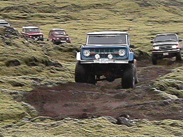 Blue early Ford Bronco with white roof leading a convoy of 4x4s across mossy off-road terrain