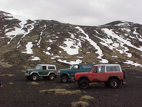 1974 Ford Bronco - Three Ford Broncos are parked on a dark, rocky landscape with snow-dusted hills in the background.