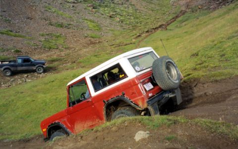 1974 Ford Bronco - A red and white Ford Bronco navigates a challenging off-road terrain, while another vehicle observes from higher ground.