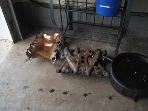 1979 Ford Bronco - Engine parts, including a rusty intake and exhaust manifold, sit on a concrete floor beside an oil drain pan.