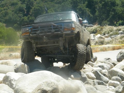 1982 Ford Bronco - This heavily modified off-road vehicle is shown tackling a challenging rocky terrain, kicking up dust as it maneuvers.