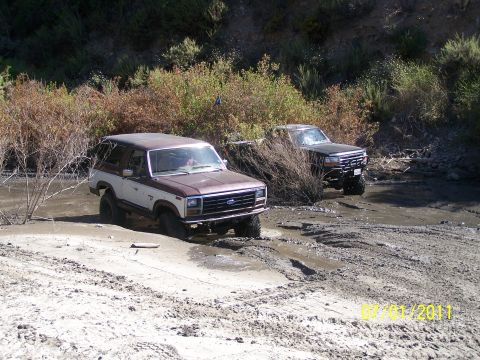 1982 Ford Bronco - Two off-road vehicles navigate a muddy terrain, with the lead vehicle appearing to be a 1980s Ford Bronco.