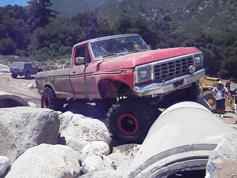 1982 Ford Bronco - A red pickup truck is shown off-roading with a yellow loader and another vehicle in the background.