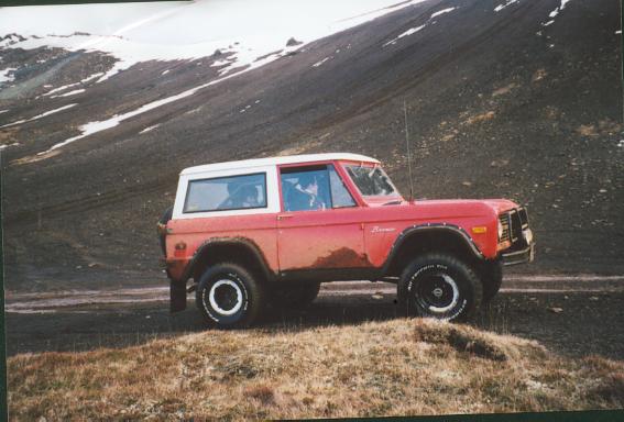 Red early Ford Bronco with white top parked on a mountain trail below snowy slopes
