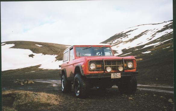 Red early Ford Bronco with white top and auxiliary lights on a gravel road beneath snow-patched mountains