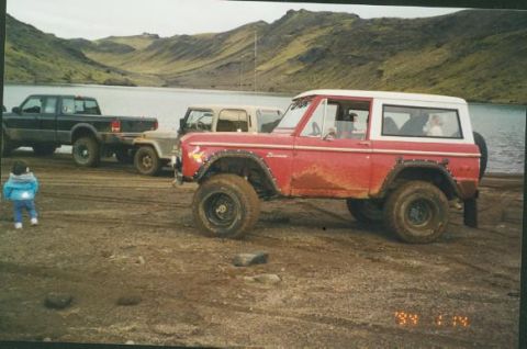 1974 Ford Bronco - Several off-road vehicles are parked near a body of water with a mountainous landscape in the background.