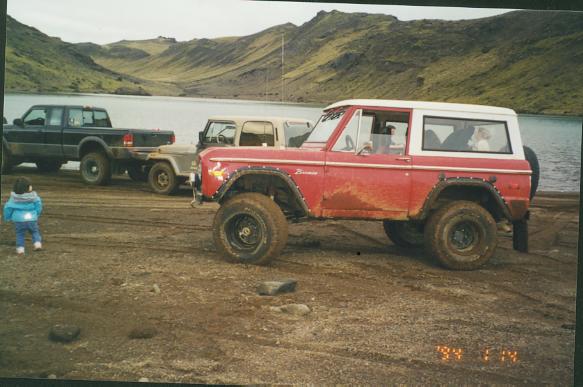 Red white-topped lifted Ford Bronco parked by a lake with mountains behind