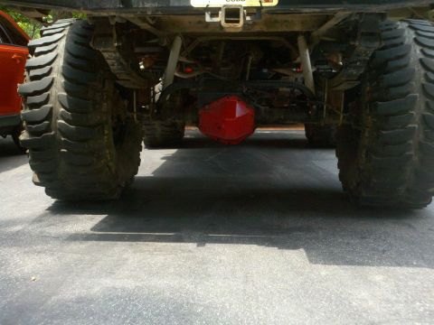 1979 Ford Bronco - The rear axle and massive tires of a heavily modified vehicle are visible, with a portion of an orange vehicle to the left.