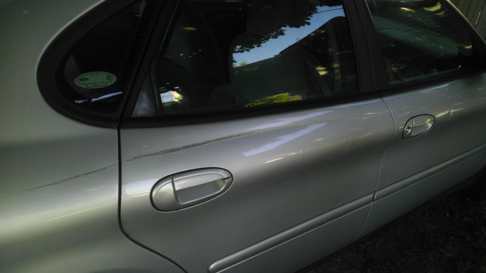 Side view of a silver sedan's rear door and window. Reflections of trees and sky are visible in the window