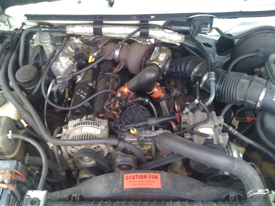 Engine bay with turbocharger, alternator, and hoses visible. Caution fan sticker on radiator shroud